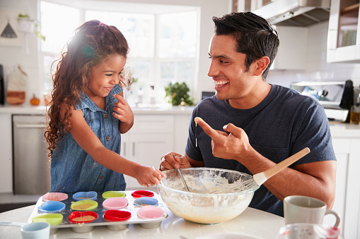 Young girl stands at the kitchen table making cakes with her father, tasting the cake mix, close up