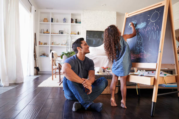 dad hispanico sentada en el piso de sala de estar viendo a su joven hija dibujo en pizarra - sentado en el suelo fotografías e imágenes de stock