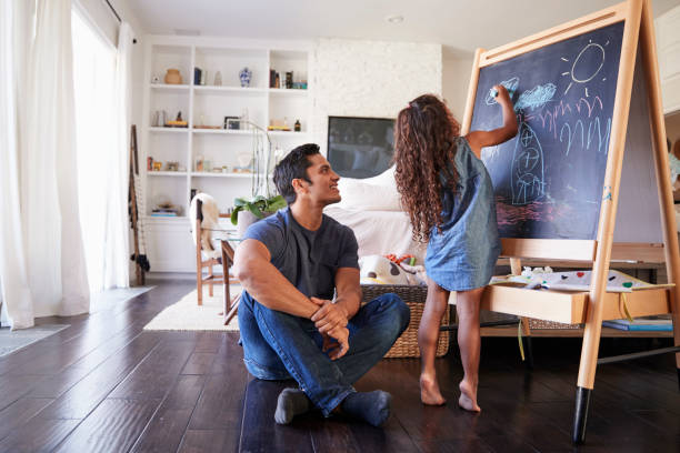 Hispanic dad sitting on the floor in sitting room watching his young daughter drawing on blackboard Hispanic dad sitting on the floor in sitting room watching his young daughter drawing on blackboard sitting on floor stock pictures, royalty-free photos & images