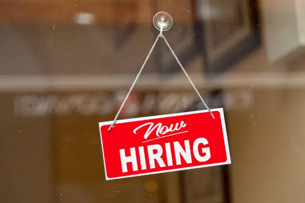 Red sign hanging at the glass door of a shop saying: "Now hiring".