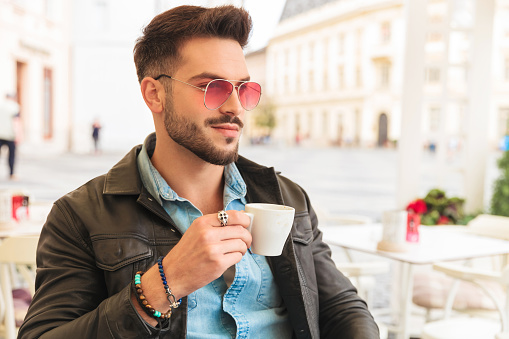 handsome man having coffee break on urban background looks to side while holding cup and sitting at table, portrait picture