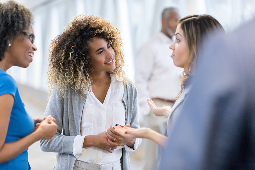 Attentive businesswomen smile while listening to a female colleague. The colleague gestures as she talk with the women.