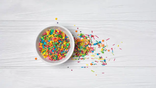 Top view on bright colorful sugar sprinkles or confetti in white bowl as baking decor on white wooden background