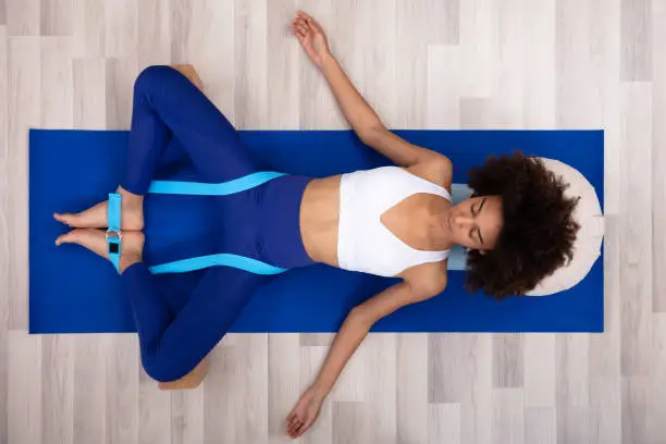 Relaxed Young Woman Lying On Fitness Mat Doing Exercise With Yoga Belt And Two Blocks