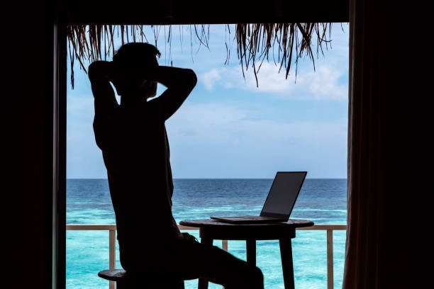 istock silhouette of a young man relaxing while working with a computer on a table. Clear blue tropical water as background 1094171232