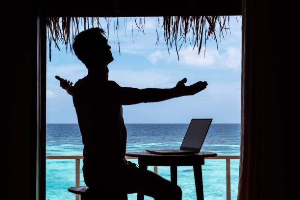 istock silhouette of a young man with arms raised working with a computer on a table. Clear blue tropical water as background 1094171260
