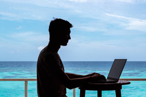 istock silhouette of a young man working with a computer on a table. Clear blue tropical water as background 1094171332