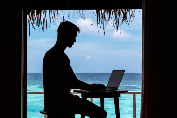 istock silhouette of a young man working with a computer on a table. Clear blue tropical water as background 1094171414