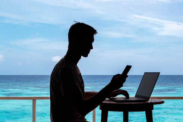 istock silhouette of a young man working with a computer and a smartphone on a table. Clear blue tropical water as background 1094171512