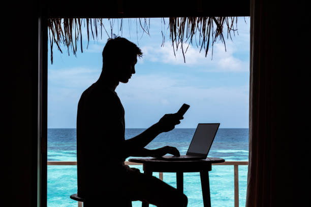 silhouette of a young man working with a computer and a smartphone on a table. Clear blue tropical water as background