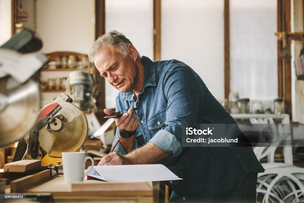 Senior carpenter working at his workshop Senior carpenter working at his workshop while standing at desk using mobile phone and writing in a book. Mature man working in carpentry workshop. Small Business Stock Photo