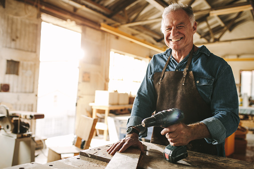 Smiling senior carpenter standing at workbench with a drill machine. Happy mature male carpenter at his workshop.