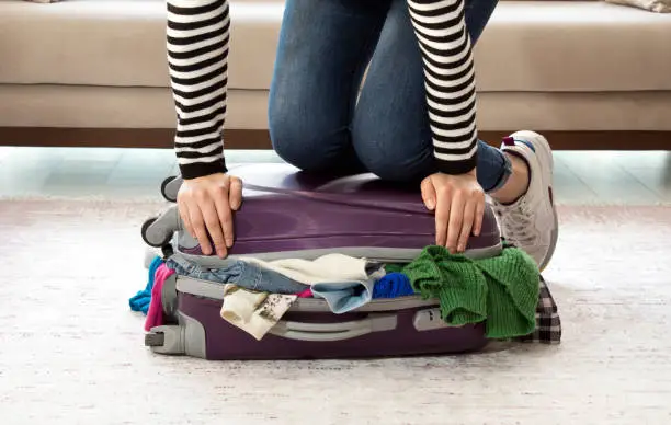 Photo of Young woman standing on her knees on overfilled suitcase, trying to close it.