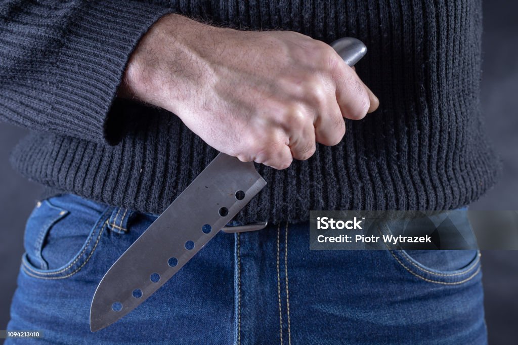 Sharp steel knife held in the hand against the background of jeans trousers. Dangerous poison in the hands of an assassin. Dark background. Adult Stock Photo