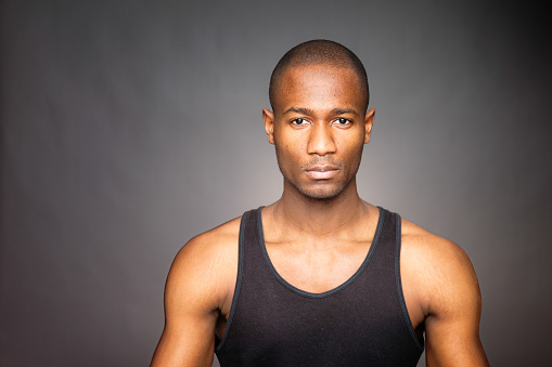 Serious Haitian man on dark background. He is wearing a tank top and looking at the camera with a troubled expression.