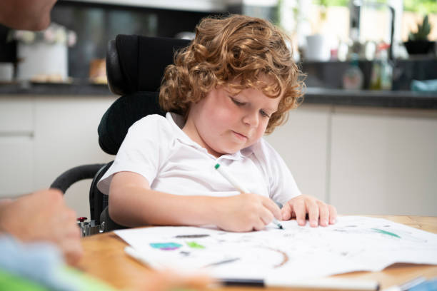Young boy in wheelchair drawing at dining table 6 year old boy with muscular dystrophy at dining table sitting in powered wheelchair, drawing on paper with crayons, and concentrating special needs child pictures stock pictures, royalty-free photos & images