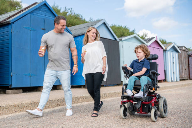 Parents smiling at son operating wheelchair 6 year old boy with muscular dystrophy in wheelchair with parents, mid adult mother and mature father, spending quality time at seaside, walking alongside their son clacton on sea stock pictures, royalty-free photos & images