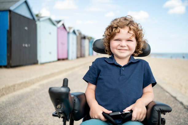 Disabled young boy sitting in wheelchair by beach huts 6 year old boy with muscular dystrophy sitting in his wheelchair, looking and smiling towards the camera on day trip to the seaside essex england photos stock pictures, royalty-free photos & images