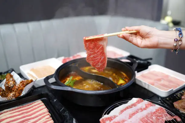 Holding raw sliced beef using chopsticks on hot pot with smoke for Japanese hot pot also known as Shabu Shabu or Sukiyaki is Japanese style.