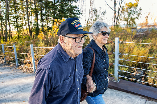 A proud, elderly, 93 year old real person senior adult man USA WWII and Korean Conflict military war veteran and his real life senior adult woman home caregiver daughter are walking side by side sightseeing on a sunny day at a public park on the shoreline of Lake Ontario. He is wearing a common, unbranded, generic souvenir shop military veteran commemorative baseball style cap with generic wording, replica plastic insignia pins and replica campaign ribbon iron-on patches. Photo taken near Rochester, N.Y., in the Finger Lakes region of western New York State, USA.\n\nNOTE: There are no official or authentic military uniform elements and no intent to portray anything other than a patriotic United States military veteran.
