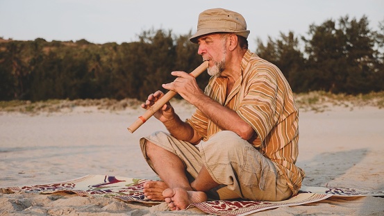 Senior man playing bamboo flute on the sandy beach next to fishing boat