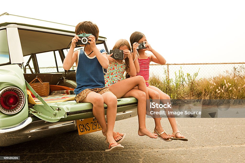 Three children sitting on back of estate car taking photographs  Retro Style Stock Photo
