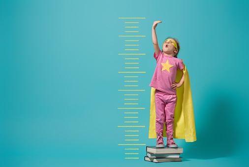 Little child playing superhero. Kid measures the growth on the background of bright blue wall. Girl power concept. Yellow, pink and  turquoise colors.