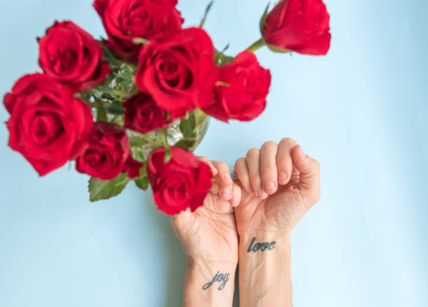 Wrist tattoos of joy and love with red roses High angle view of wrists with joy and love tattoos and defocused red roses on blue background - valentines day concept (selective focus) wrist tattoo stock pictures, royalty-free photos & images