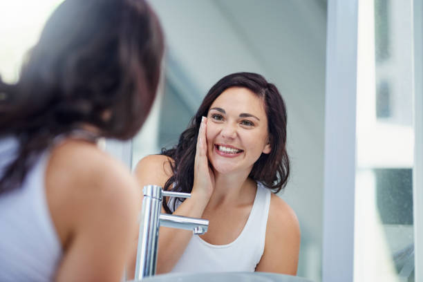 That glow that says you're good to go Shot of an attractive young woman admiring her face in the bathroom mirror looking in mirror stock pictures, royalty-free photos & images