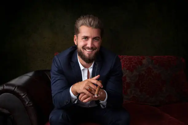 Portrait of handsome businessman in elegant suit sitting on couch and holding a cigar