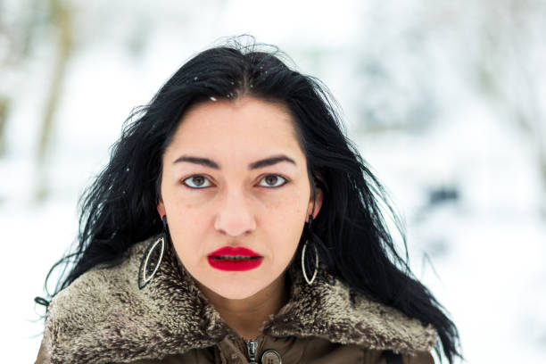 Close up color image depicting the portrait of a young brunette woman in a snowy park in winter. The bright white snow is blurred in the background. Focus is on the woman's face. She has a serious and moody expression on her face, and she is wearing bright red lipstick. The woman is in her mid 30s. Room for copy space.