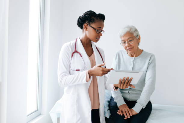 Black female doctor showing digital tablet to senior patient A black female doctor standing next to female patient and showing her something on digital tablet. doctor stock pictures, royalty-free photos & images