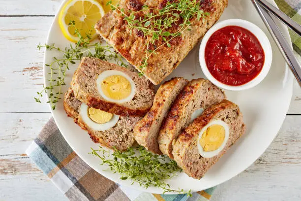sliced Meatloaf stuffed with hard-boiled egg served with tomato sauce and  lemon slices on a white plate with fork and knife on an old wooden table, view from above, close-up, flatlay