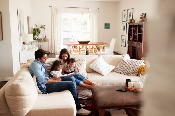 young hispanic family sitting on sofa reading a book together in the living room, seen from doorway - education relaxation women home interior imagens e fotografias de stock