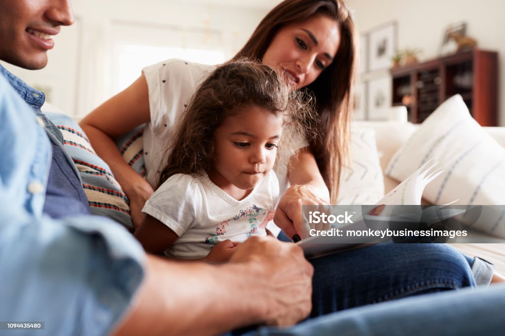 Hispanic couple and their young daughter sitting on the sofa reading a book together at home Family Stock Photo