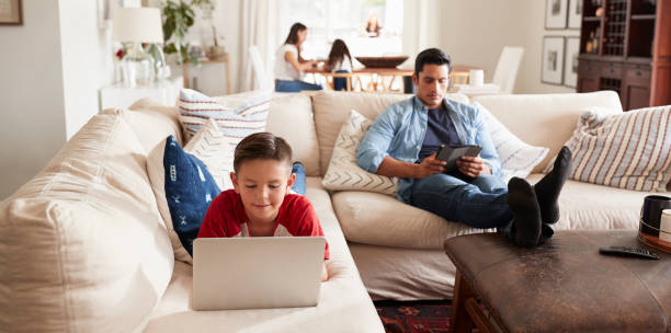 pre-teen junge auf sofa mit laptop, papa sitzen mit tablet, mutter und schwester im hintergrund - wohnanlage stock-fotos und bilder
