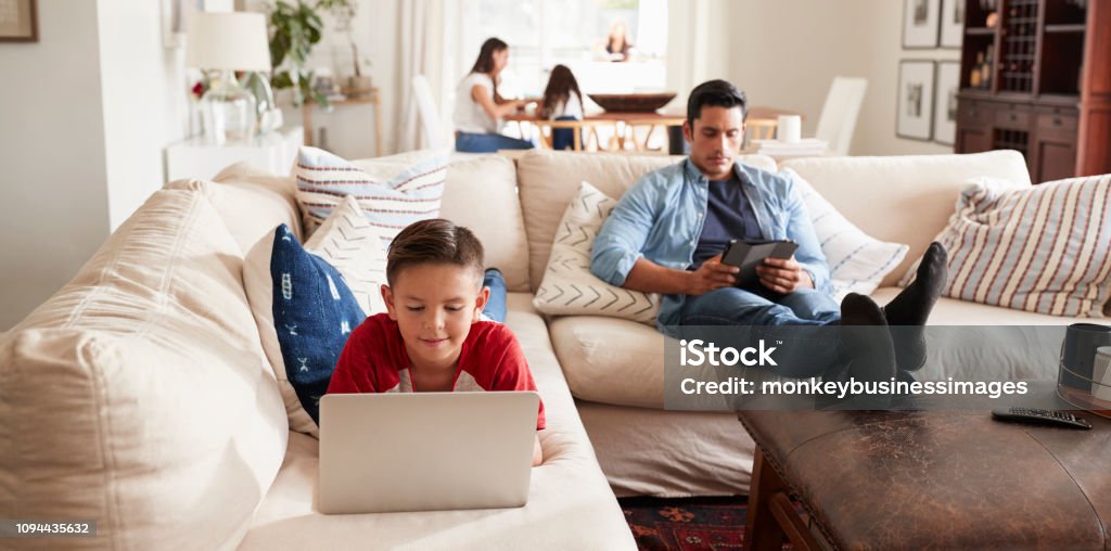 Pre-teen boy lying on sofa using laptop, dad sitting with tablet, mum and sister in the background Family Stock Photo