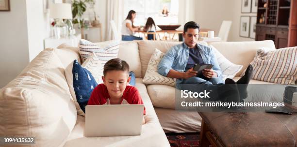 Preteen Boy Lying On Sofa Using Laptop Dad Sitting With Tablet Mum And Sister In The Background Stock Photo - Download Image Now