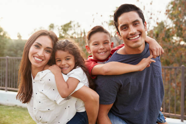 Young Hispanic parents piggyback their children in the park, smiling to camera, focus on foreground Young Hispanic parents piggyback their children in the park, smiling to camera, focus on foreground four people photos stock pictures, royalty-free photos & images