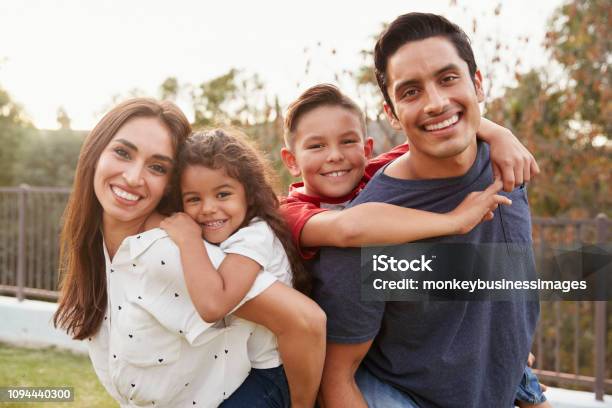 Young Hispanic Parents Piggyback Their Children In The Park Smiling To Camera Focus On Foreground Stock Photo - Download Image Now