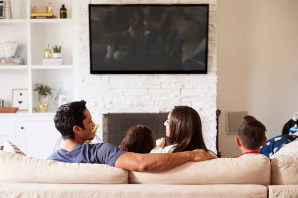 vista posterior de la familia hispana de cuatro sentada en el sofá viendo la televisión, mamá mira papá - family television watching watching tv fotografías e imágenes de stock