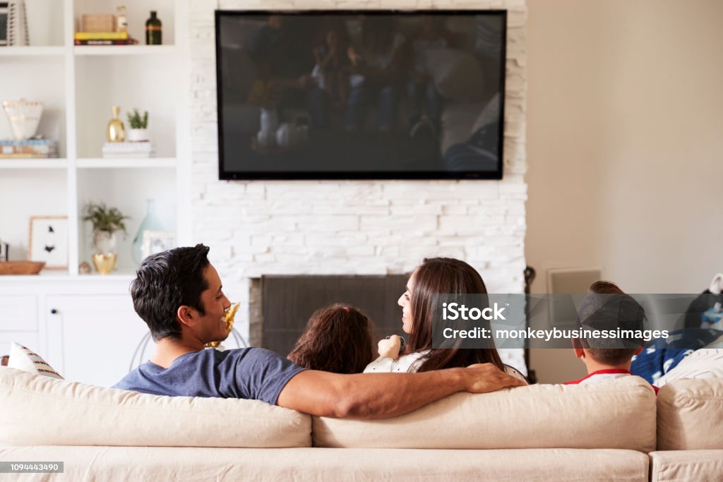 Back view of young Hispanic family of four sitting on the sofa watching TV, mum looking at dad Family Stock Photo