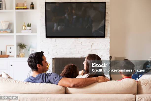 Back View Of Young Hispanic Family Of Four Sitting On The Sofa Watching Tv Mum Looking At Dad Stock Photo - Download Image Now