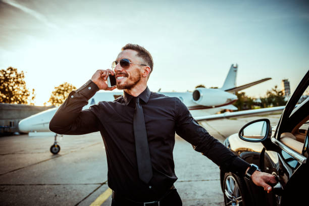 istock Young rich man smiling while talking on a mobile phone while opening a car door next to a parked private airplane 1094443514