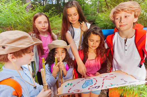 Group of kids in forest examine and point with finger on map during treasure hunting hike activity in summer camp