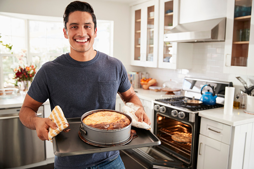 Smiling millennial Hispanic man standing in kitchen presenting the cake he has baked to camera