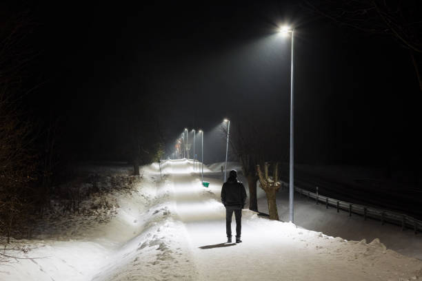 istock Young man alone slowly walking on snow covered sidewalk under white street lights. Peaceful atmosphere in snowy night. Back view. 1094445308