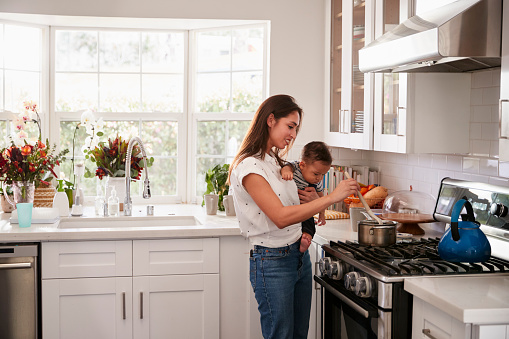 Multitasking mum holding her young baby while she makes food at the hob in her kitchen, side view