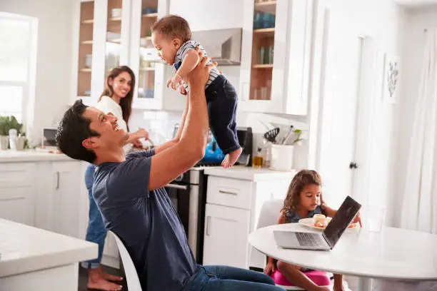 Photo of Young Hispanic family in their kitchen, dad lifting baby in the air, mum cooking at hob, close up