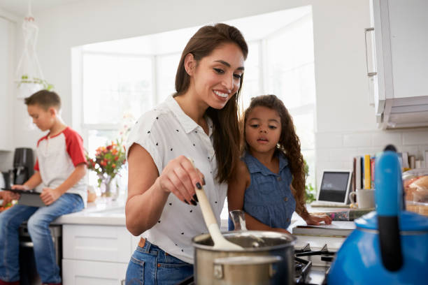 madre e giovane figlia che preparano il cibo al piano cottura in cucina, figlio pre-adolescente seduto sullo sfondo - sibling baby three people baby girls foto e immagini stock